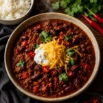 A bowl of homemade Chilli Con Carne with beef mince, kidney beans, and red peppers in a rich spiced tomato sauce, topped with soured cream, grated cheese, and fresh coriander, served with white rice