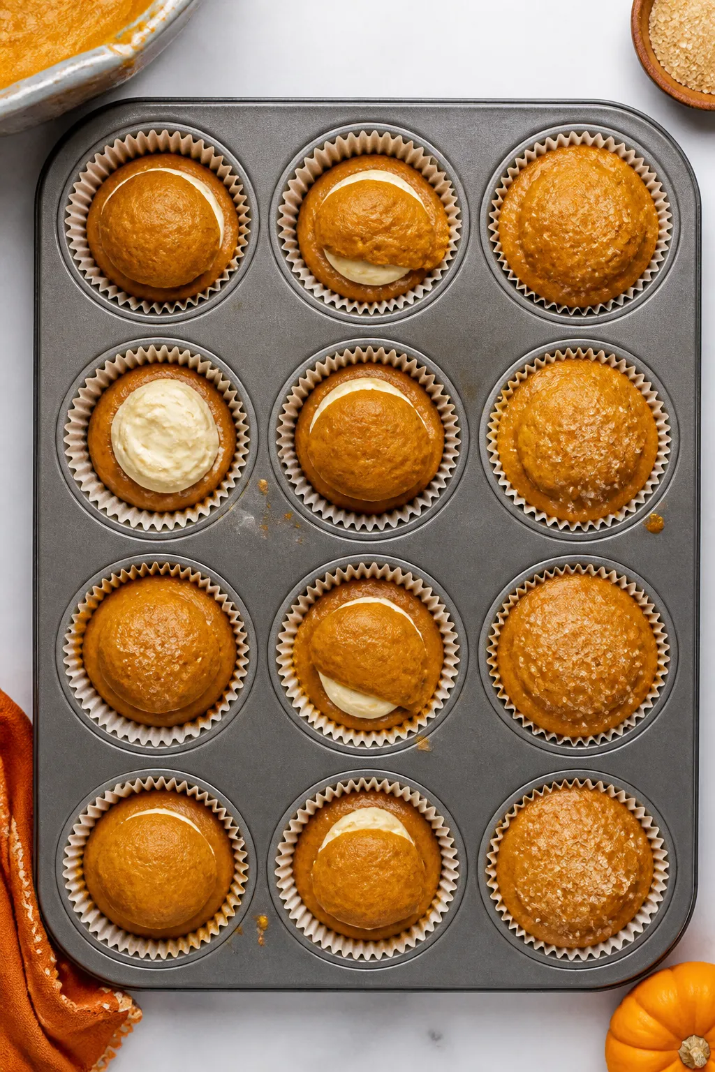 Overhead process shot of a lined 12-cup muffin tin during assembly: lower layer of pumpkin batter pr